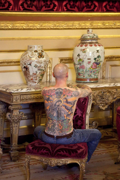 Tim Steiner exposant son tatouage parmi les autres oeuvres, assis de dos, écoutant de la musique sur un siège qui est aussi son socle au Louvre en 2012.
