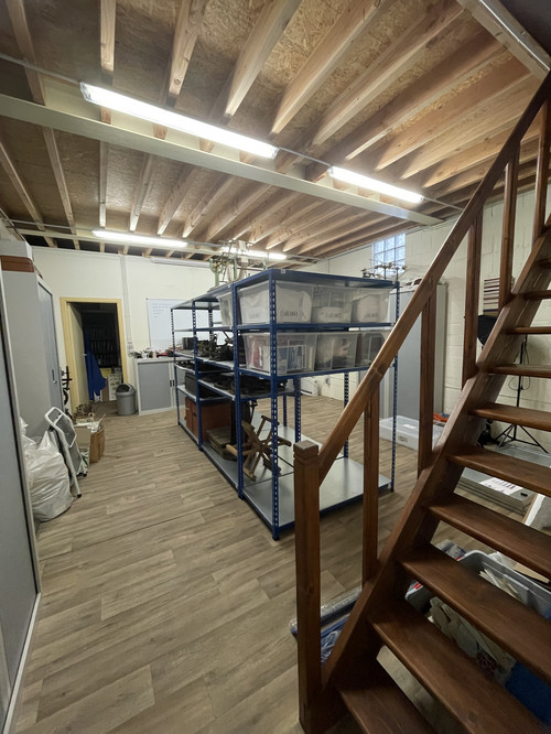 La même salle après réorganisation. Les objets industriels soigneusement rangés sur des rayonnages métalliques bleus et dans des boîtes transparentes. L’espace apparaît désormais ordonné et optimisé pour le stockage des collections. 